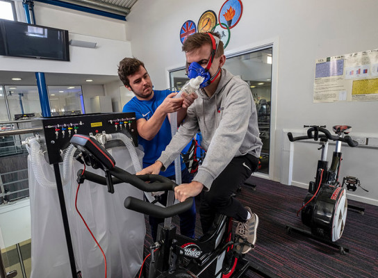 Student supporting another student on exercise bike in Paston gym